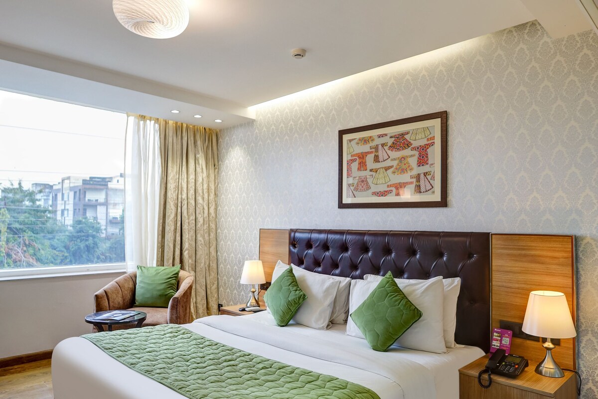 A hotel room with a large bed, a chair, a bedside table with a lamp, and a view of the city.