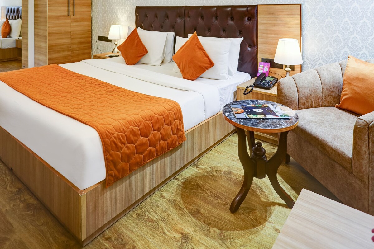 A hotel room with a large bed, a sofa, a small round table, and a nightstand with a lamp.