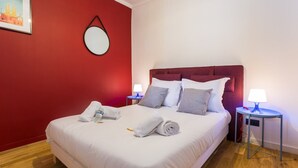 A neatly made bed with white linens and rolled towels, a red accent wall, a round mirror, and two bedside lamps on a wooden floor. - Cosy apartment - 4P - Place de la Bastille (Paris)