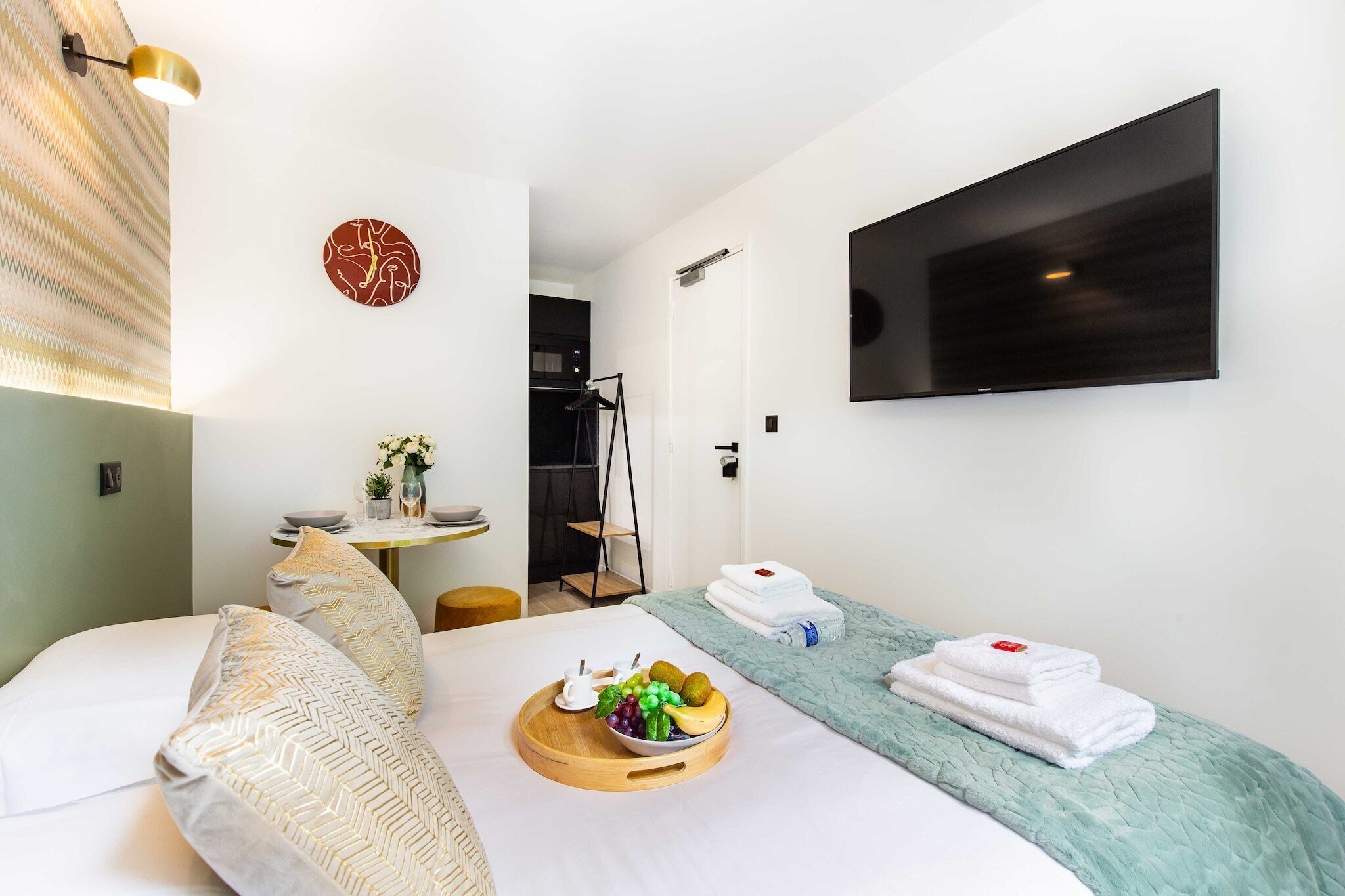 Una camera d'albergo moderna con un letto, un piccolo tavolo con frutta, una TV e un angolo doccia.