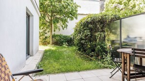 Un patio con mesa y sillas, un pequeño jardín con césped y plantas, y un edificio blanco con ventana y puerta.