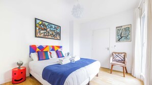 Un dormitorio moderno con cama, almohadas coloridas, una mesita de noche roja y obras de arte en las paredes.