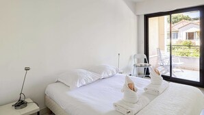 Ein Hotelzimmer mit Bett, weißen Kissen, einem Nachttisch mit Lampe und Blick auf einen Balkon mit Gartenmöbeln.