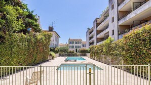 Ein Poolbereich mit rechteckigem Pool, umgeben von einem weißen Metallzaun, und angrenzende Gebäude mit Balkonen.