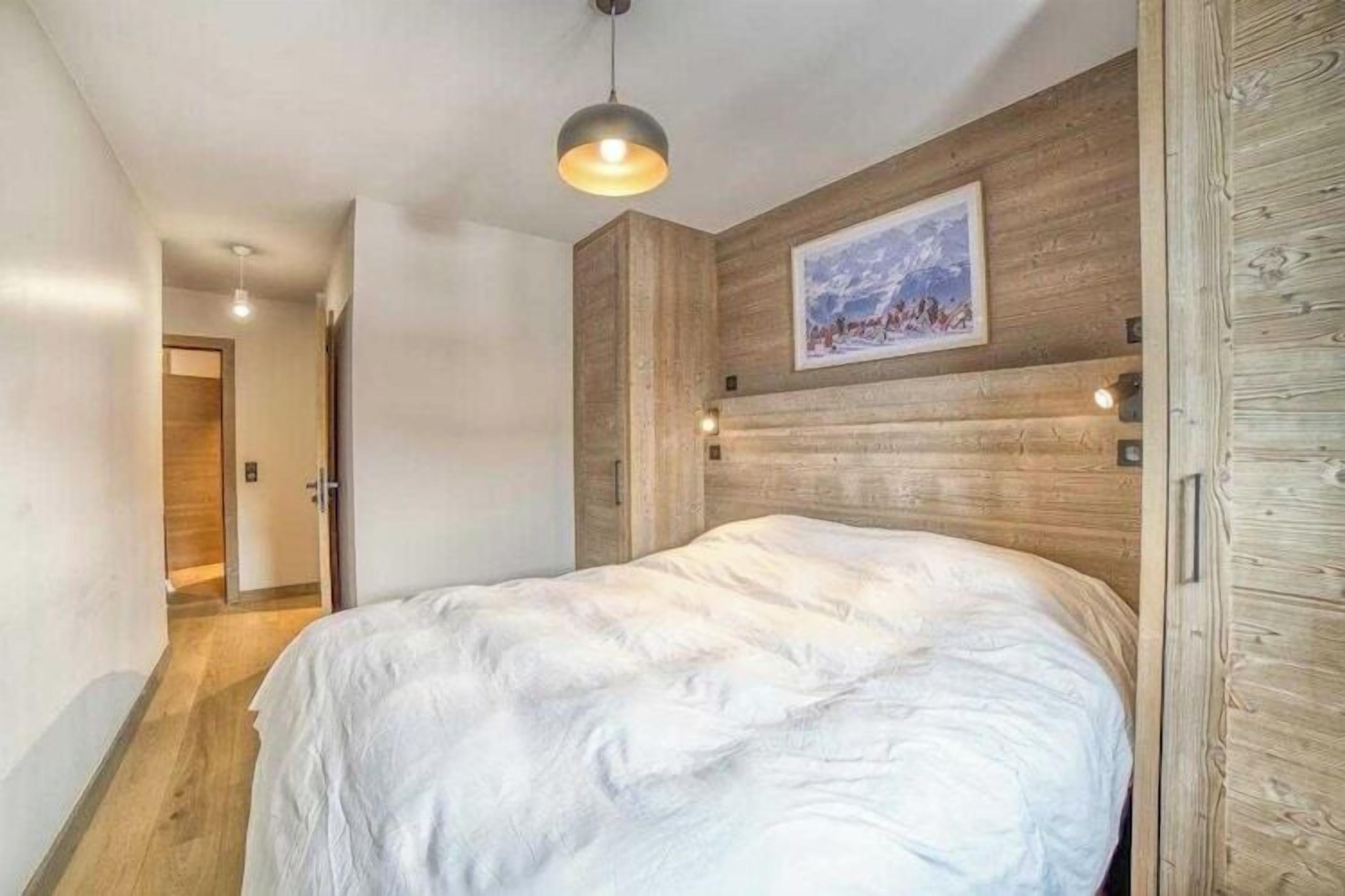 A bedroom with a wooden headboard, a bed with white bedding, a pendant light, and a painting on the wall.