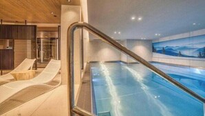 An indoor swimming pool with a glass barrier, lounge chairs, and a view of mountains. - Luxurious 4-room apartment with swimming pool, gym, spa and parking (Megève)