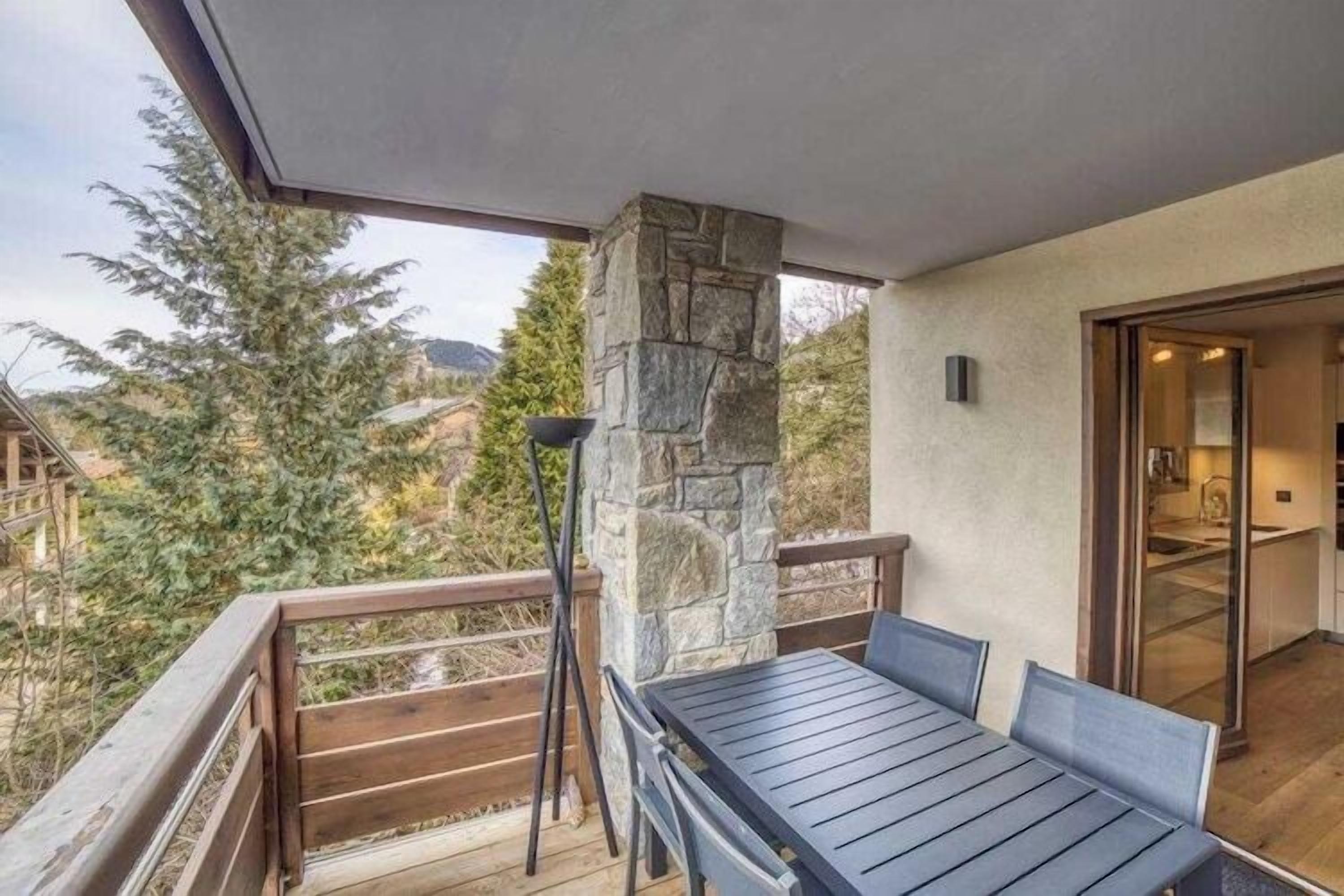 A balcony with a stone pillar, a table with chairs, and a view of trees and houses.