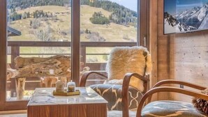 A room with a large window offering a view of a hilly landscape. There is a wooden table with a decorative item on it, and a chair with a fur throw. - Luxurious 4-room apartment with swimming pool, gym, spa and parking (Megève)
