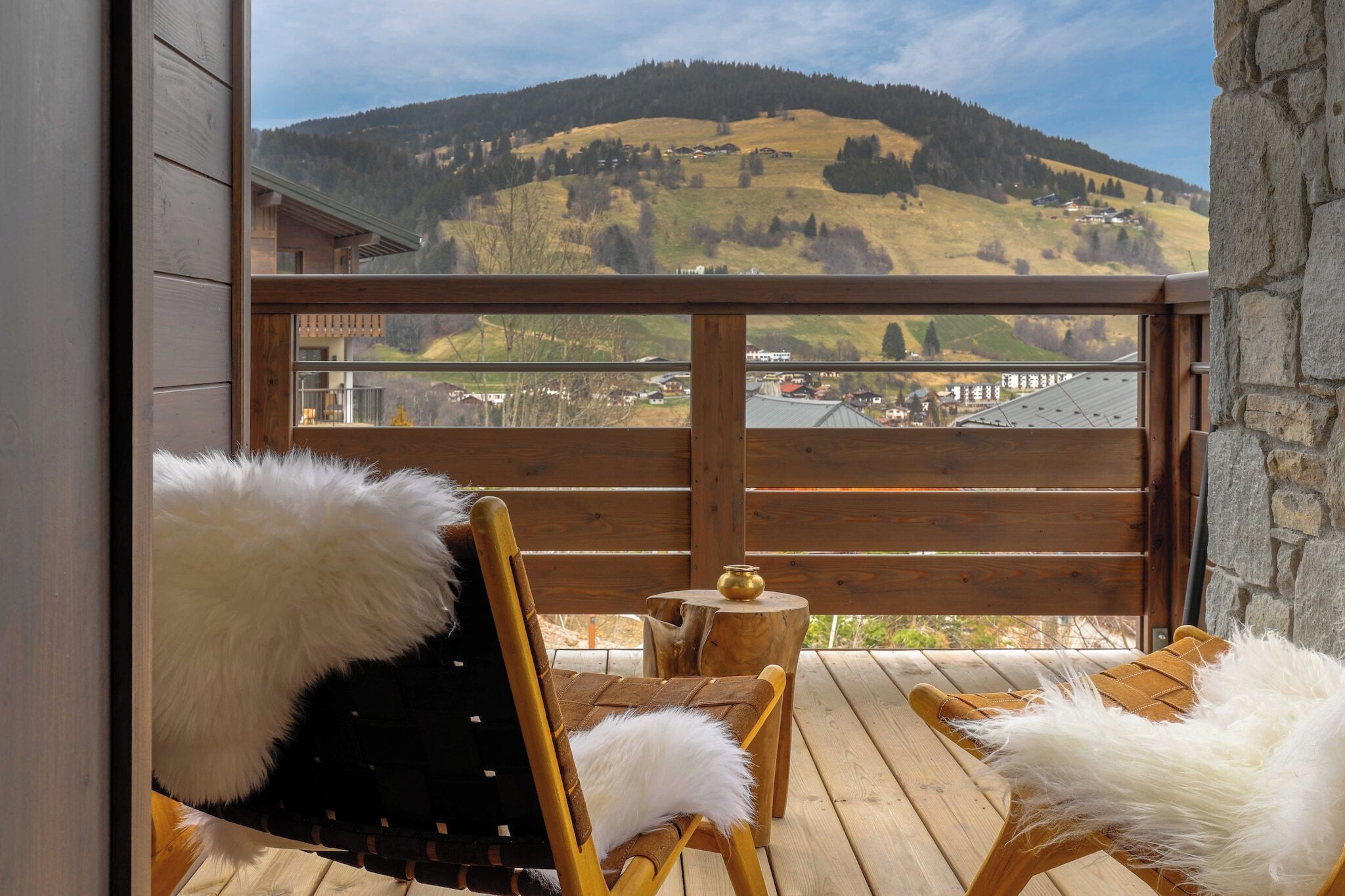 Une terrasse en bois avec deux chaises, chacune agrémentée d’un coussin moelleux blanc, donnant sur un paysage montagneux avec un village en contrebas.
