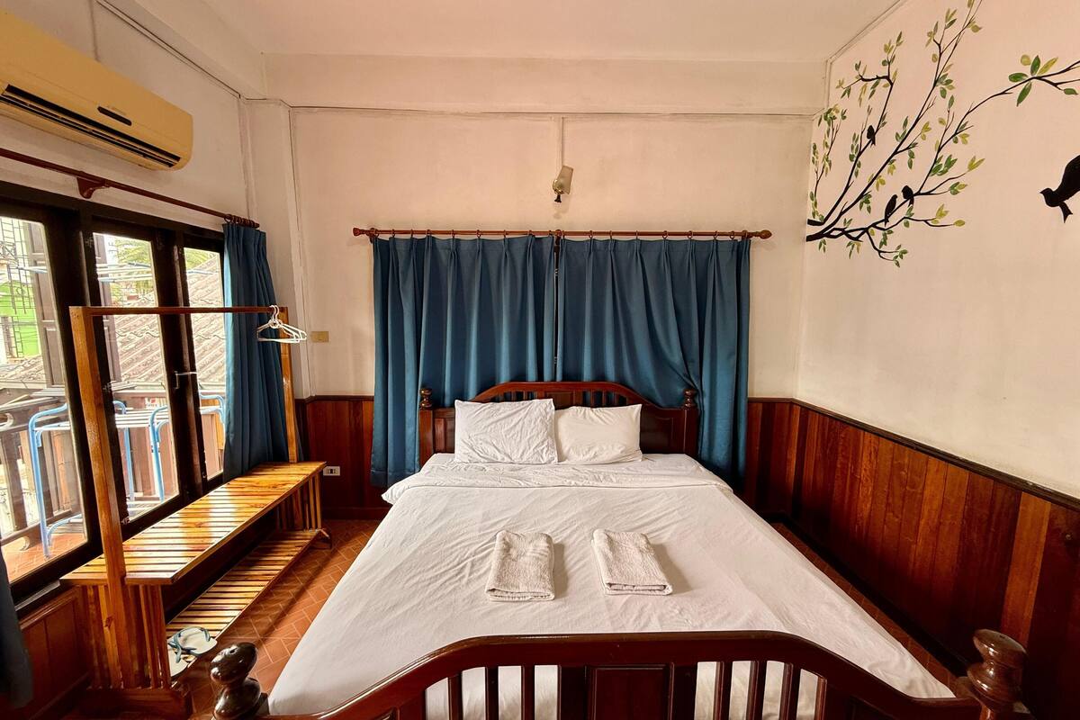A bedroom with a wooden bed, white linens, a view through a window, and a wall-mounted air conditioner.