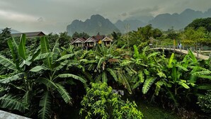A lush garden with banana plants and a bridge leading to a row of wooden houses with red roofs. - Domon Riverside Hotel (Vang Vieng)