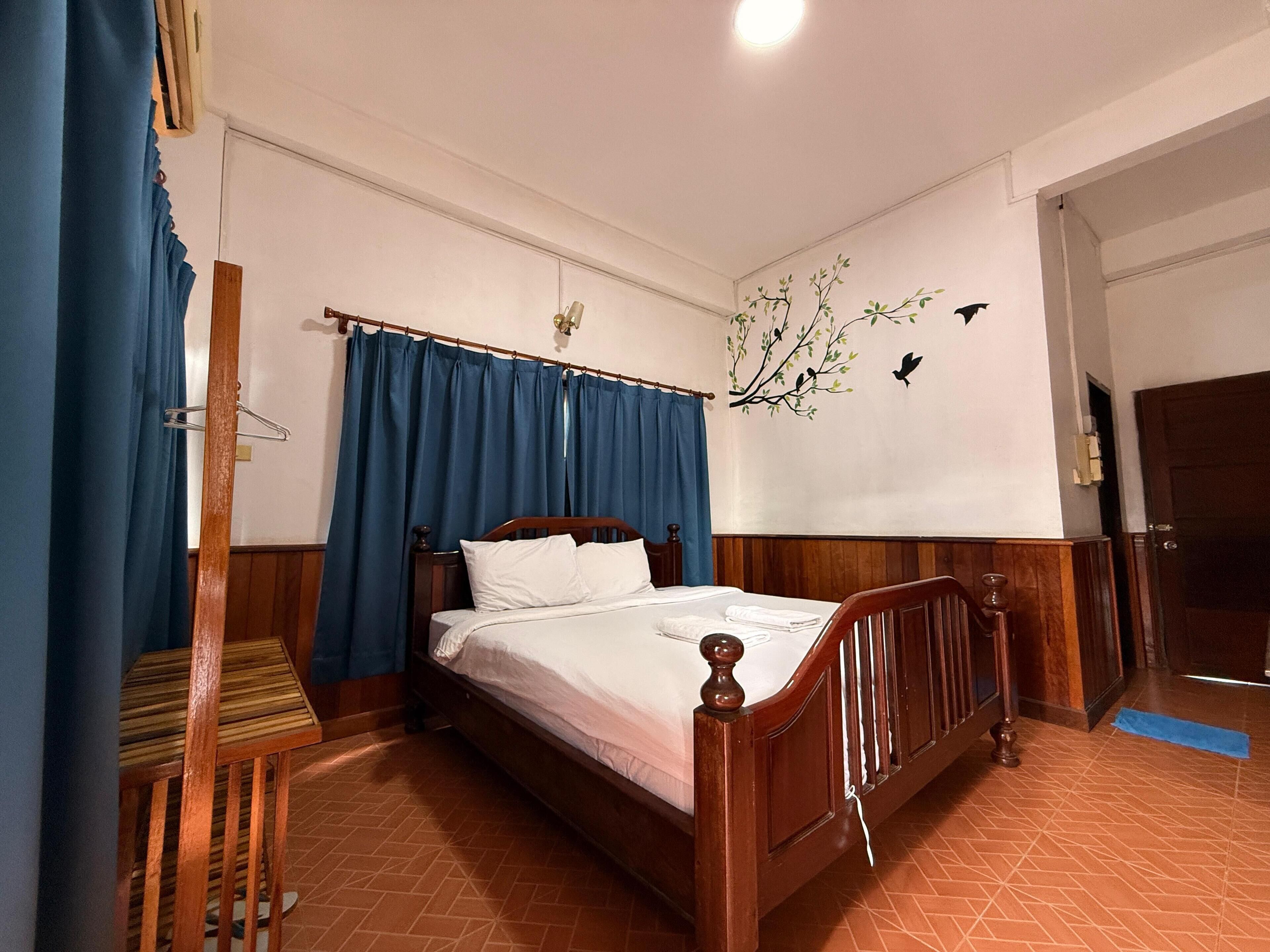 A bedroom with a wooden bed, blue curtains, a wooden chair, and a wall with a bird mural.