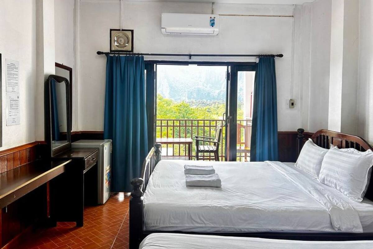 A hotel room with two beds, a desk, a chair, and a balcony with a view of greenery.