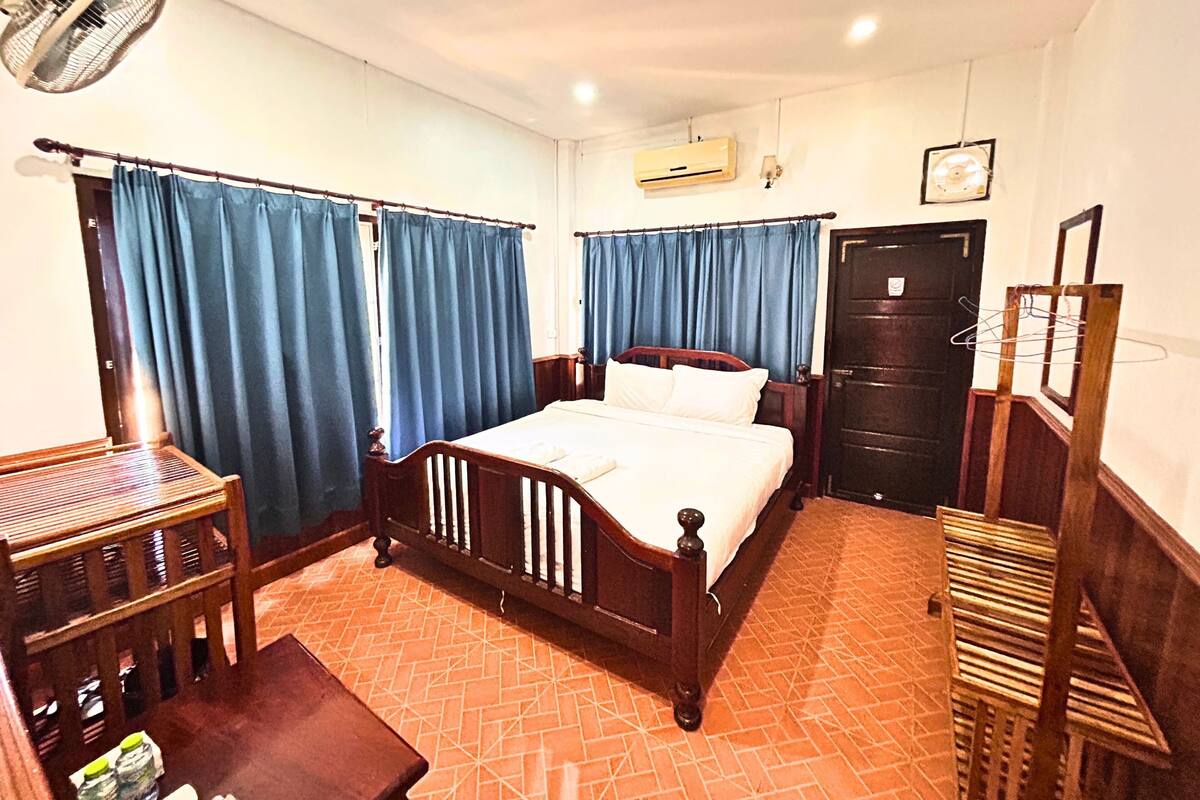 A bedroom with a wooden bed, blue curtains, an air conditioning unit, a fan, a door, and a small table with bottles.