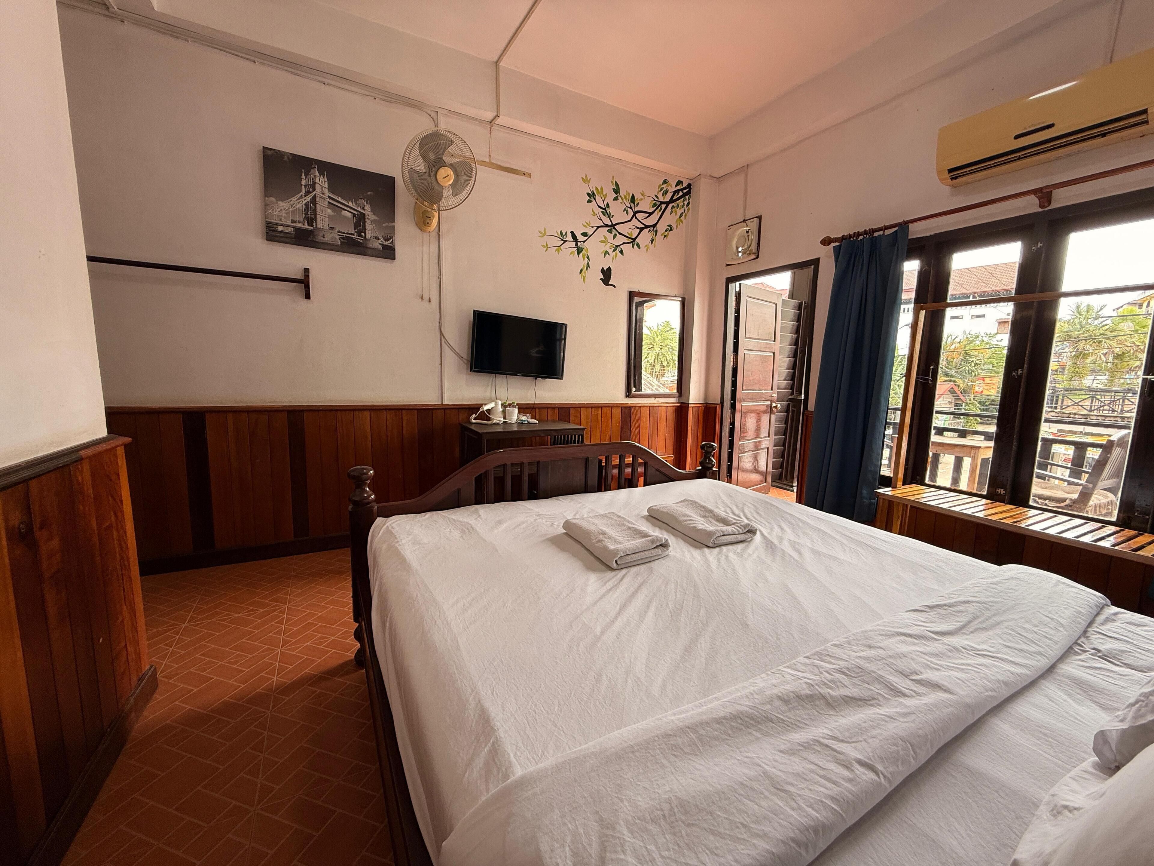 Une chambre d’hôtel équipée d’un lit, d’une télévision, d’un ventilateur et d’une fenêtre donnant sur l’extérieur.