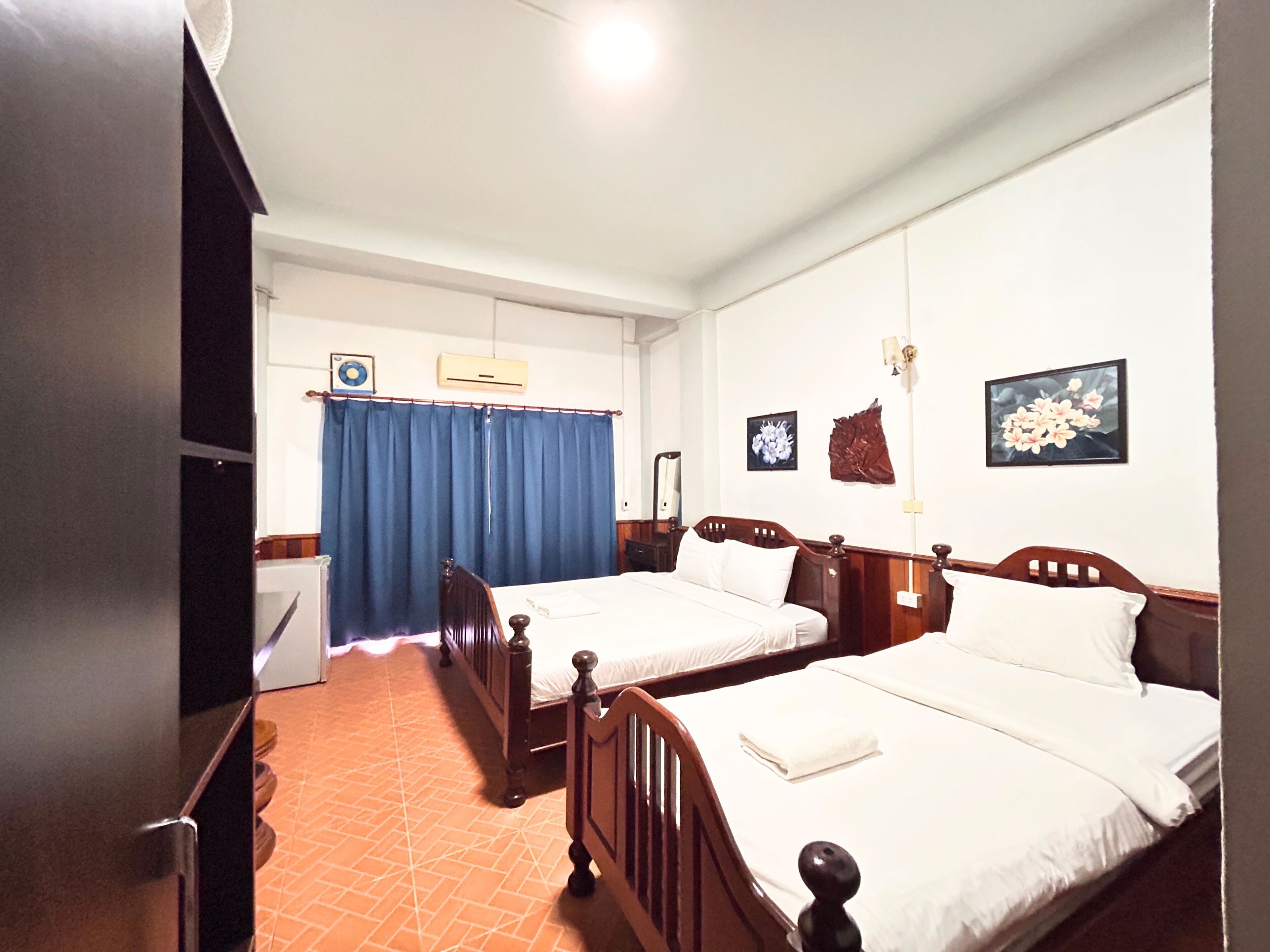 Una camera d'albergo con due letti di legno, una tenda blu, un condizionatore d'aria a parete e un tavolino con una lampada.