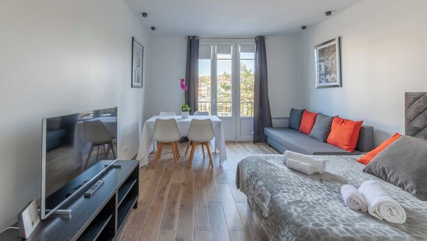 A modern hotel room with a flat-screen TV, a dining table with chairs, a sofa with orange pillows, and a bed with rolled towels.