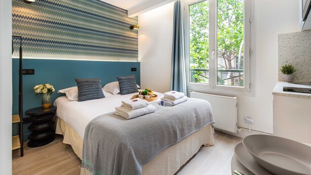 A modern hotel room with a large bed, a view of greenery through a window, a small kitchen area, and a decorative wall pattern. - Cosy Studio - Beaugrenelle parc des expositions 4 (Paris)