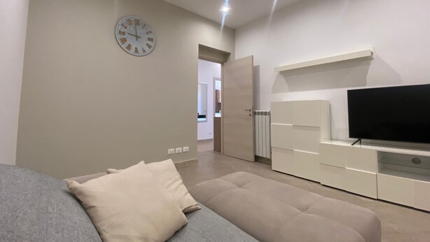 Een moderne woonkamer met een grijze bank, een beige voetenbankje, een flatscreen-tv op een wit kastje, en een wandklok.