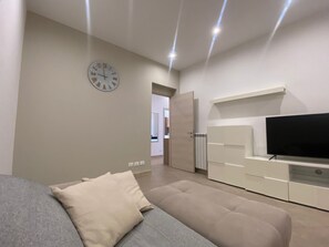 Un salon moderne avec un canapé gris, un pouf beige, un téléviseur à écran plat posé sur un meuble blanc et une horloge murale.