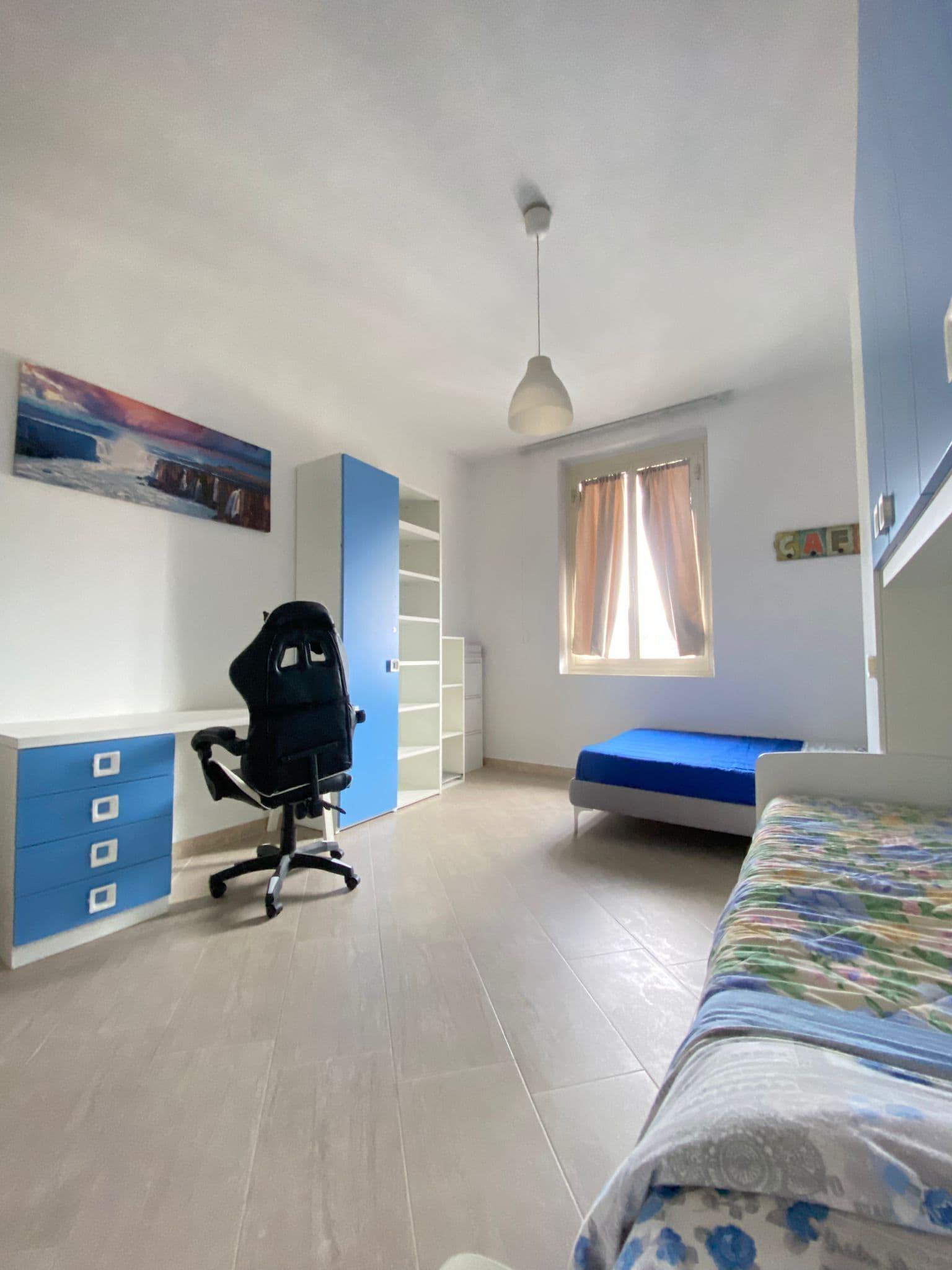 Une chambre à coucher avec un lit, un bureau doté de tiroirs, une chaise, une fenêtre avec des rideaux et un tableau accroché au mur.