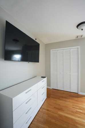 Een slaapkamer met een wandtelevisie, een witte dressoir en een inloopkast met schuifdeuren.