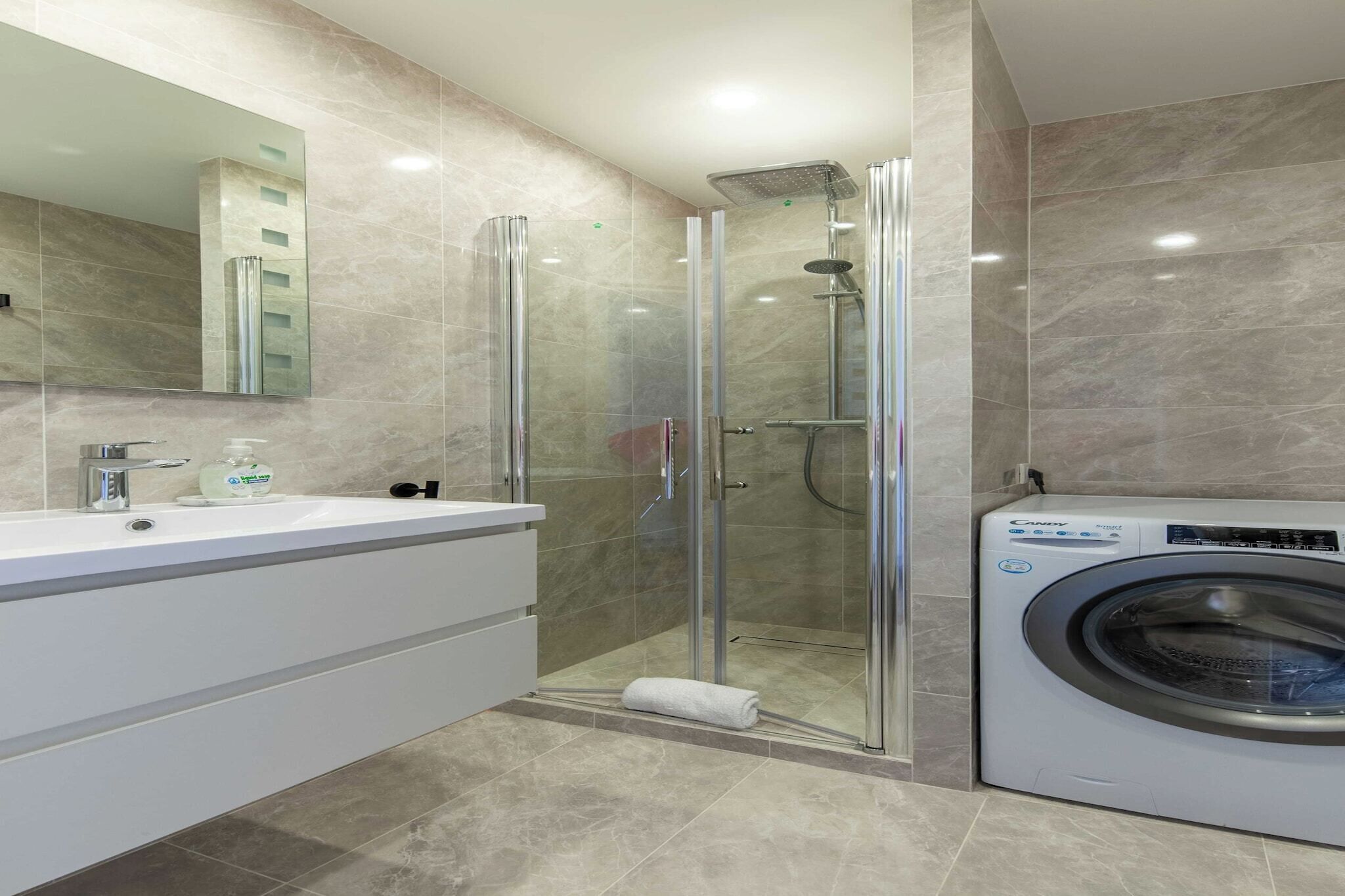 Un baño moderno con ducha de vidrio, un lavamanos blanco con espejo y una lavadora de carga frontal.
