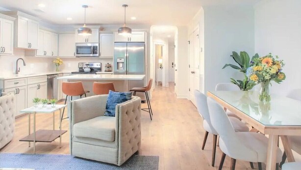 Una cocina moderna con gabinetes blancos, electrodomésticos de acero inoxidable e iluminación colgante. Hay un comedor con mesa y sillas de vidrio, y una sala de estar con sofá capitonado y una pequeña mesa de centro.