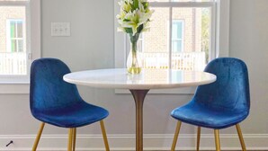 A room with a round white table, two blue chairs with gold legs, and a vase with flowers on the table.
