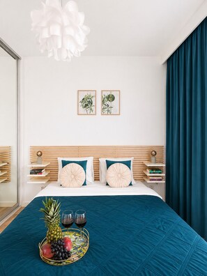 Une chambre d’hôtel avec un lit, une tête de lit, une table de chevet, un plateau avec des fruits et des boissons, et une grande fenêtre avec des rideaux.