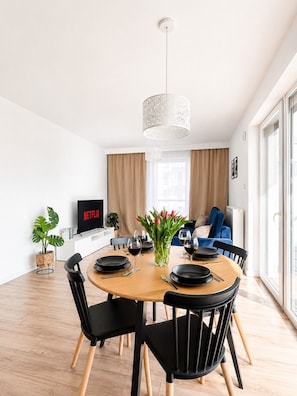 Un salon moderne comprenant un coin repas, un téléviseur diffusant Netflix, une plante et un vase avec des fleurs.