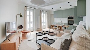 A modern living room with a sofa, coffee table, and TV. There is a dining area with chairs and a table. The kitchen is visible with cabinets and appliances. - Authentic apartment - 3BR/8P - Clignancourt (Paris)