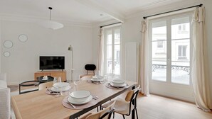 A dining area with a wooden table set for four, a television, and a view of the street through large windows. - Authentic apartment - 3BR/8P - Clignancourt (Paris)