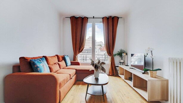 A modern living room with a sofa, a coffee table, a TV on a wooden stand, and a view of buildings through large windows with curtains.