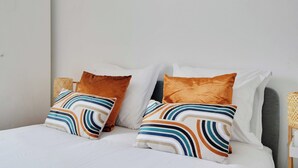 A neatly made bed with white linens and striped pillows, a grey headboard, and a woven side table.