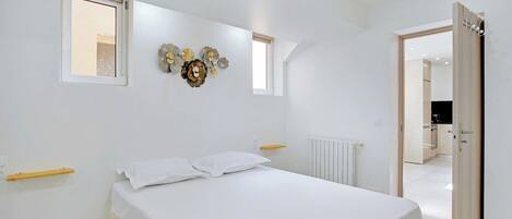 A bedroom with a bed, a window, a radiator, and a door leading to a kitchen.