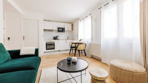 A modern living room with a green sofa, a round black coffee table, and a wicker ottoman. A kitchen with white cabinets and a dining area with a table and chairs is visible in the background. - Comfortable Studio for 2 people - Puteaux (Puteaux)