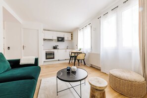 Una sala moderna con un sofá verde, una mesa de centro redonda negra y un otoman de mimbre. Se ve una cocina con armarios blancos y una zona de comedor con una mesa y sillas al fondo.