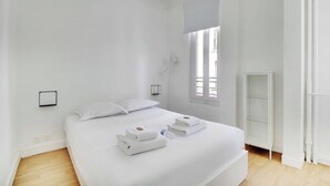 Un dormitorio minimalista con una cama grande, ropa de cama blanca, una mesita de noche, un pequeño estante y una ventana con persianas.