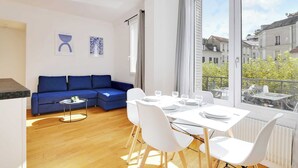 Una sala de estar moderna con un sofá azul, una mesa de comedor lista para cuatro comensales y un ventanal con vistas a la ciudad.