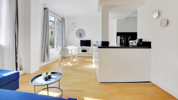 Una sala de estar moderna y bien iluminada con una mesa de comedor, sillas, un sofá azul, una pequeña mesa redonda con plantas y una cocina con encimera y electrodomésticos.