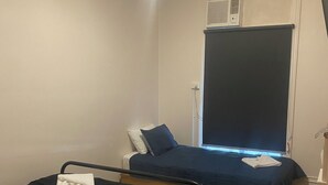 A small, dimly lit room with two single beds, each with a black mattress and white pillows. There is a window with closed blinds, and a ceiling light is visible. - Culcairn Hotel (Culcairn)