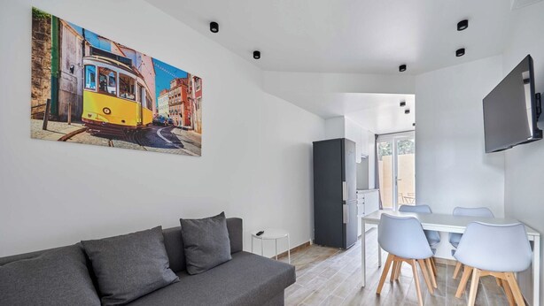 A modern living room with a gray sofa, a dining table with chairs, a flat-screen TV mounted on the wall, and a painting of a tram on the wall.