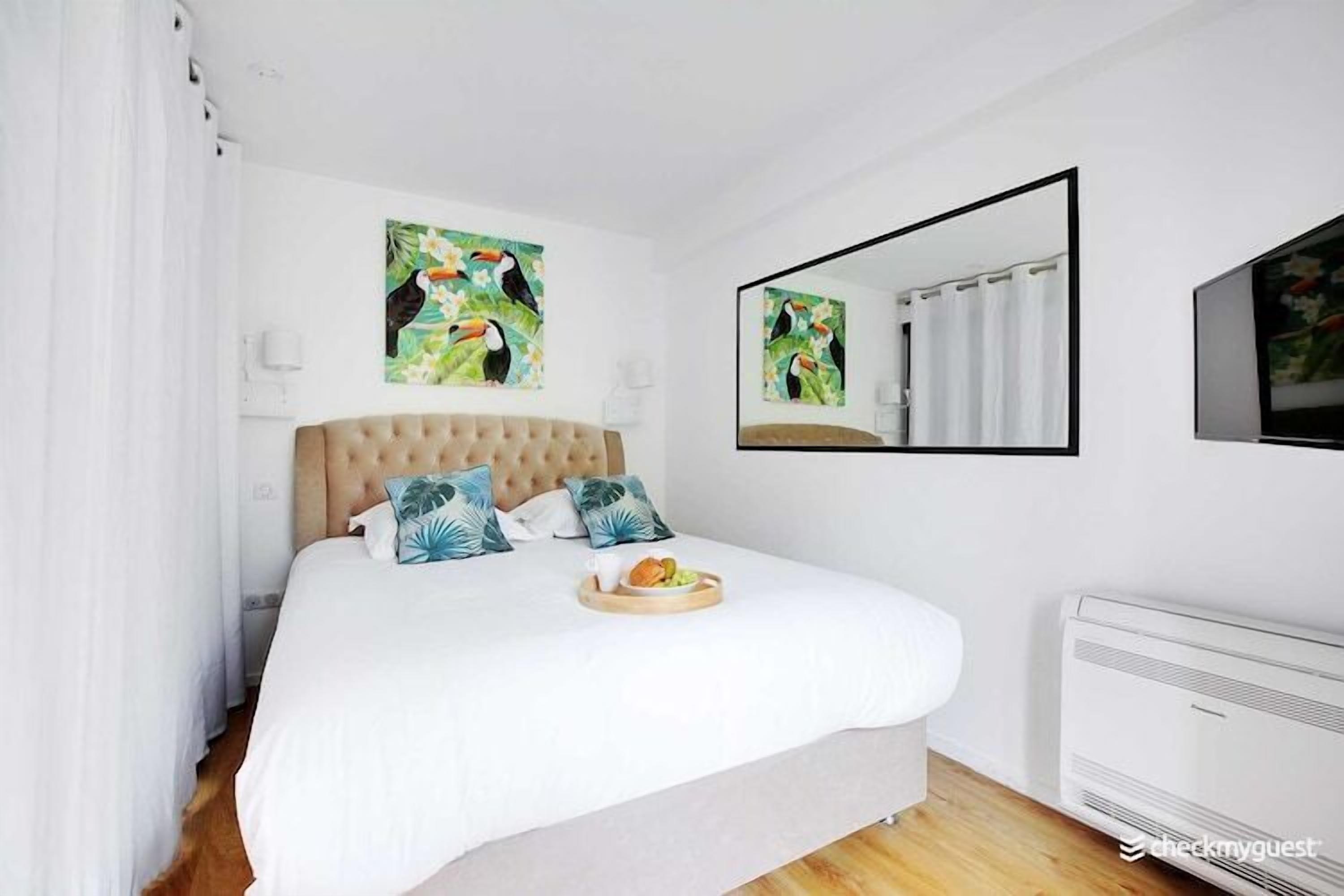 Ein Schlafzimmer mit einem großen Bett, weißer Bettwäsche, einem Frühstücksbrettchen, einem Spiegel und Kunstwerken mit tropischem Motiv.