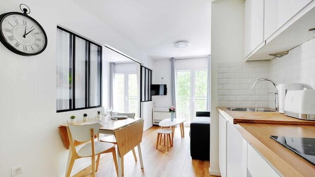 A modern kitchen with white cabinets, a wooden countertop, and a built-in oven. There is a dining area with a white table and chairs, and a large window providing natural light. - Cozy apartment - 1BR/4P Montmartre / Sacré Cœur (Paris)