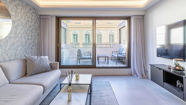 Un salon moderne avec un canapé, une table basse et un téléviseur. Il y a un balcon avec vue sur les immeubles et une table avec des chaises.