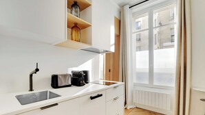 Una cocina moderna con encimera blanca, lavabo integrado y cafetera. Hay una ventana con cortinas y vistas a edificios.
