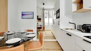 Una cocina moderna con armarios blancos, encimera negra y horno empotrado. Hay un comedor con una mesa preparada para cuatro personas y vistas a la ciudad a través de amplias ventanas.