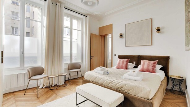 Una habitación de hotel moderna con una cama grande, una zona de estar con una silla y una mesita, y vistas de la ciudad a través de amplios ventanales.