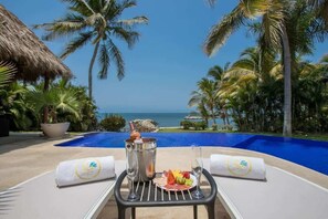 Escena junto a la piscina con vista al océano, una mesa con una botella de champán, un plato de fruta y una copa de vino.